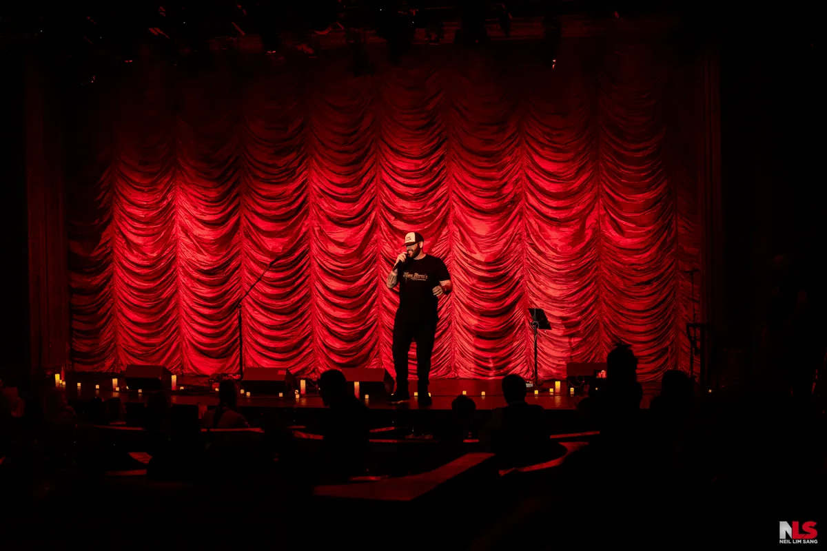 Wide shot of host on a red curtain stage with audience silhouettes in the foreground.